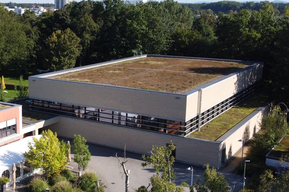 Luftaufnahme eines modernen Geb&auml;udes mit begr&uuml;ntem Flachdach, umgeben von B&auml;umen und einem Parkplatz.