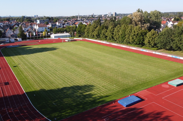 Luftaufnahme eines Sportplatzes mit gr&uuml;nem Rasen, umgeben von einer Laufbahn und B&auml;umen im Hintergrund.