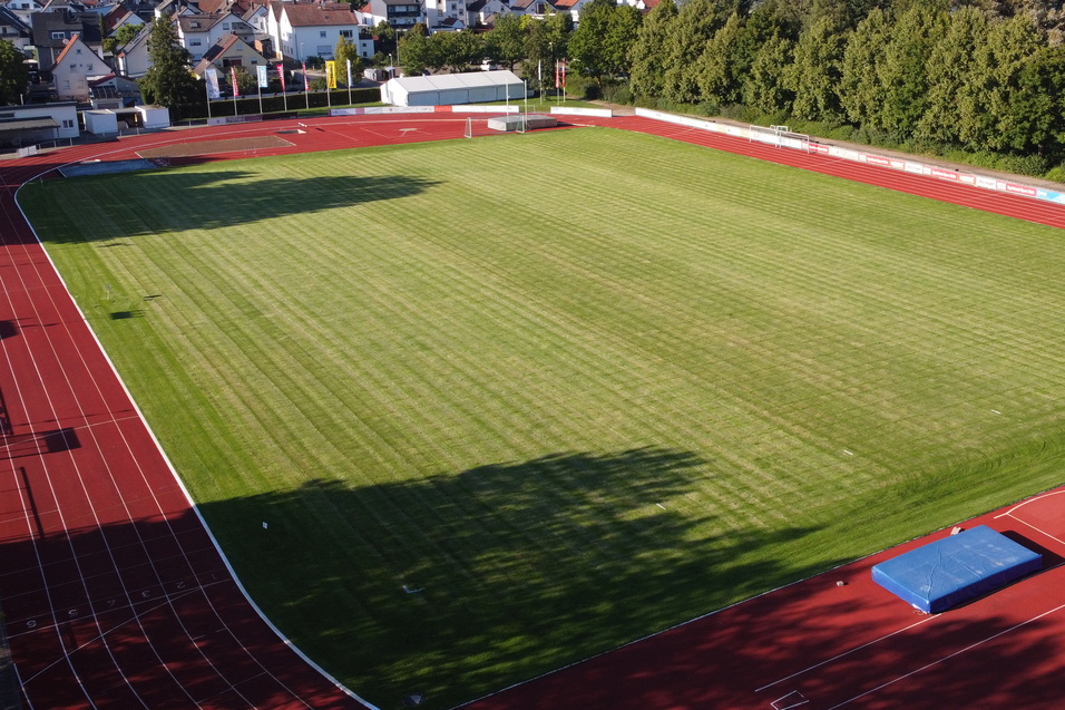 Blick auf ein Sportfeld mit gr&uuml;ner Rasenfl&auml;che und rotem Laufbahnrand, umgeben von B&auml;umen und Wohngeb&auml;uden.