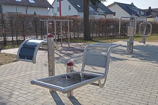 Fitnessger&auml;te im Freien auf einem gepflasterten Platz mit Wohnh&auml;usern im Hintergrund.