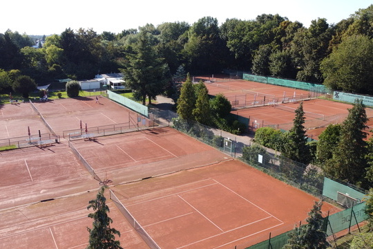 Luftaufnahme eines Tennisplatzes mit mehreren roten Sandpl&auml;tzen und umgebender Vegetation.