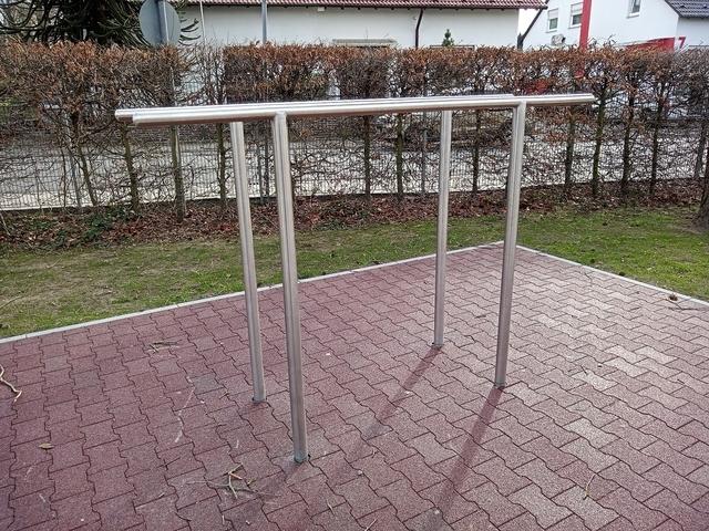 Metallene Trainingsstange auf einem roten Gummiboden in einem Park, umgeben von niedrigen Hecken.