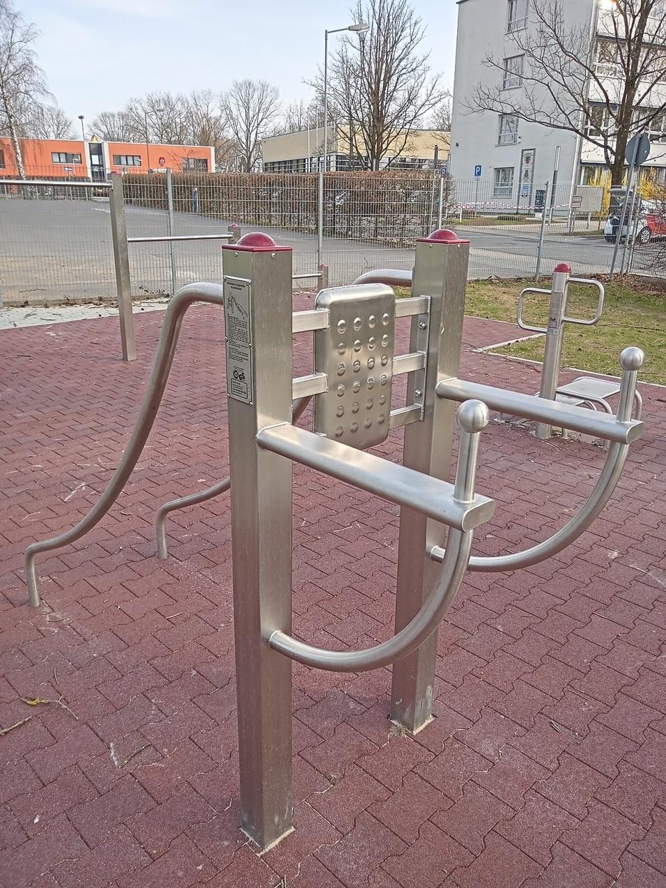 Fitnessger&auml;t im Freien mit zwei Haltegriffen auf einem roten Gummibelag, umgeben von B&auml;umen und Geb&auml;uden im Hintergrund.