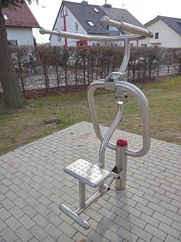 Fitnessger&auml;t im Freien mit Sitz, Hebeln und rutschfester Plattform auf einem gepflasterten Boden.