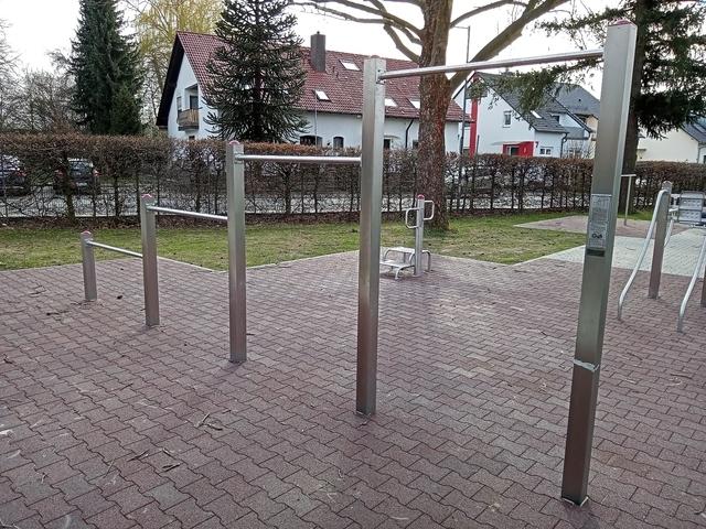 Fitness-Ger&auml;te aus Metall in einem Park, umgeben von Rasen und B&auml;umen, mit Wohnh&auml;usern im Hintergrund.