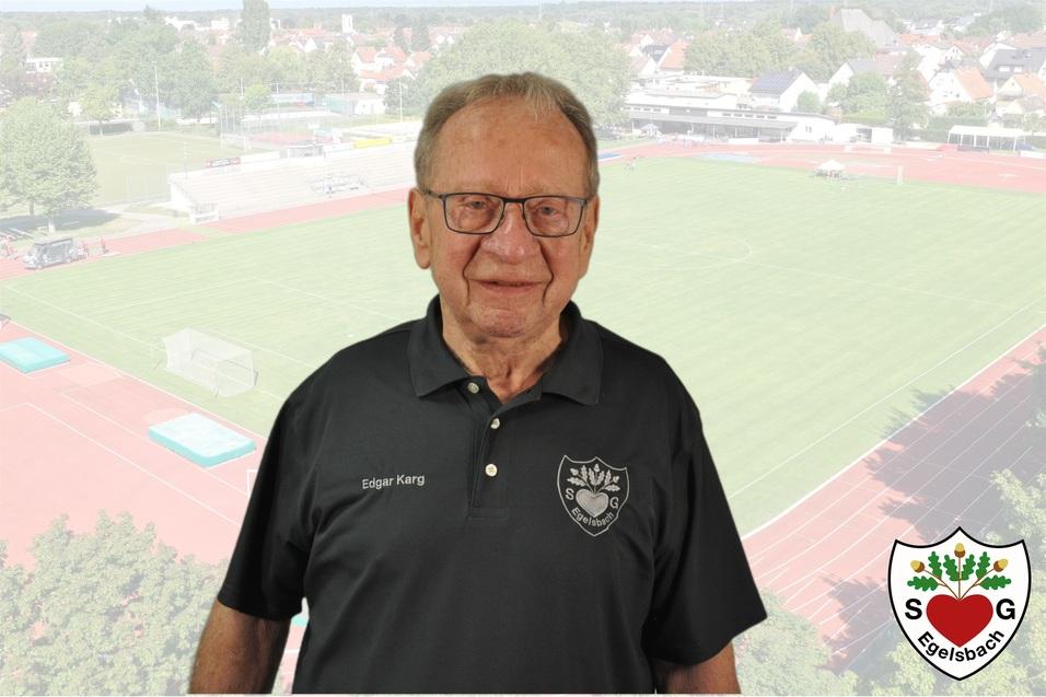 &Auml;lterer Mann mit Brille in schwarzer Sportkleidung vor einem Sportplatz mit Rasen und Laufbahn.
