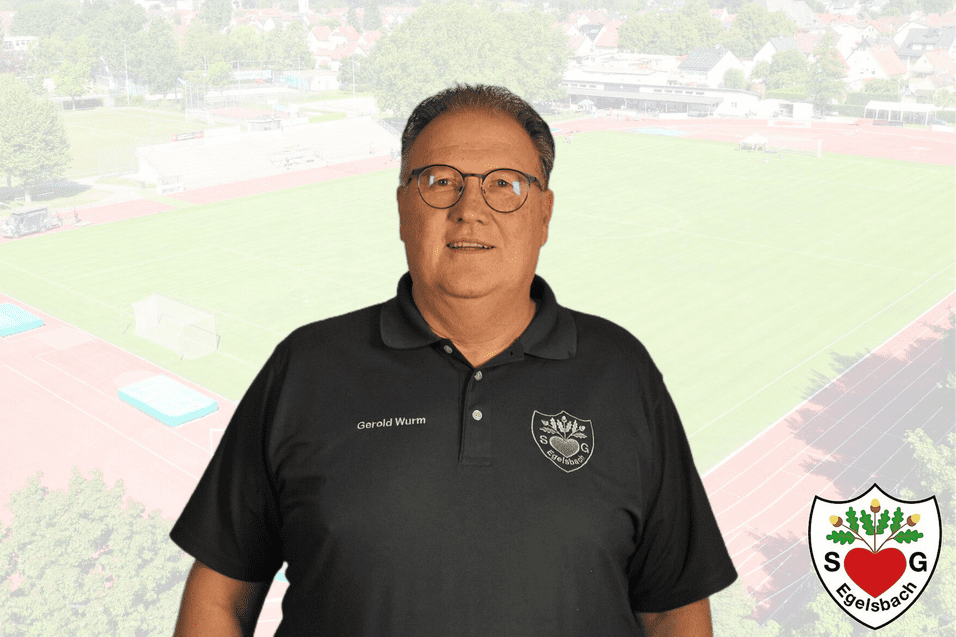 Portr&auml;t eines Mannes in schwarzer Polo-Shirt mit Vereinslogo vor einem Sportplatz-Hintergrund.