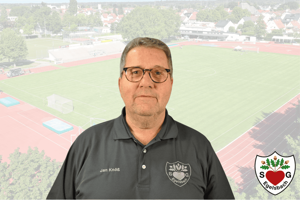 Portr&auml;t eines Mannes mit Brille im Vordergrund, Fu&szlig;ballfeld und Sportanlagen im Hintergrund. Tr&auml;gt ein dunkles Poloshirt.