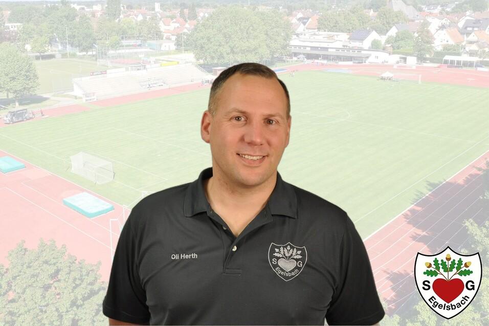 Portr&auml;t eines Mannes in Schwarz, vor einem Sportplatz mit Laufbahn und Spielfeld im Hintergrund. Logo unten rechts.