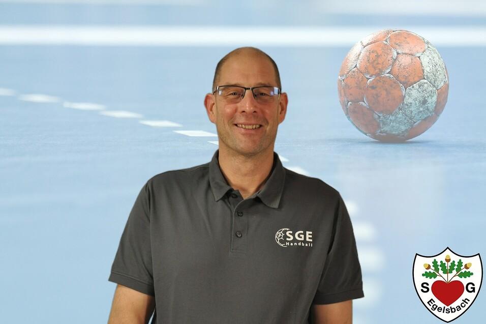 Mann in grauem Shirt lächelt vor einem Handballfeld mit Ball im Hintergrund. Logo der SGE Egelsbach unten rechts.