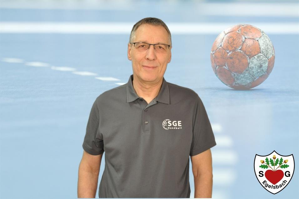Mann in grauem Poloshirt steht lächelnd vor einer verschwommenen Handballörtlichkeit mit ball.