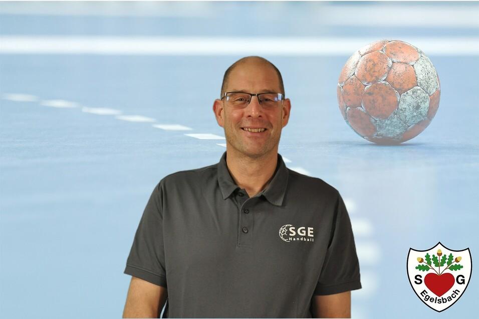 Mann in grauem Shirt l&auml;chelt vor einem Handballfeld mit Ball im Hintergrund. Logo der SGE Egelsbach unten rechts.