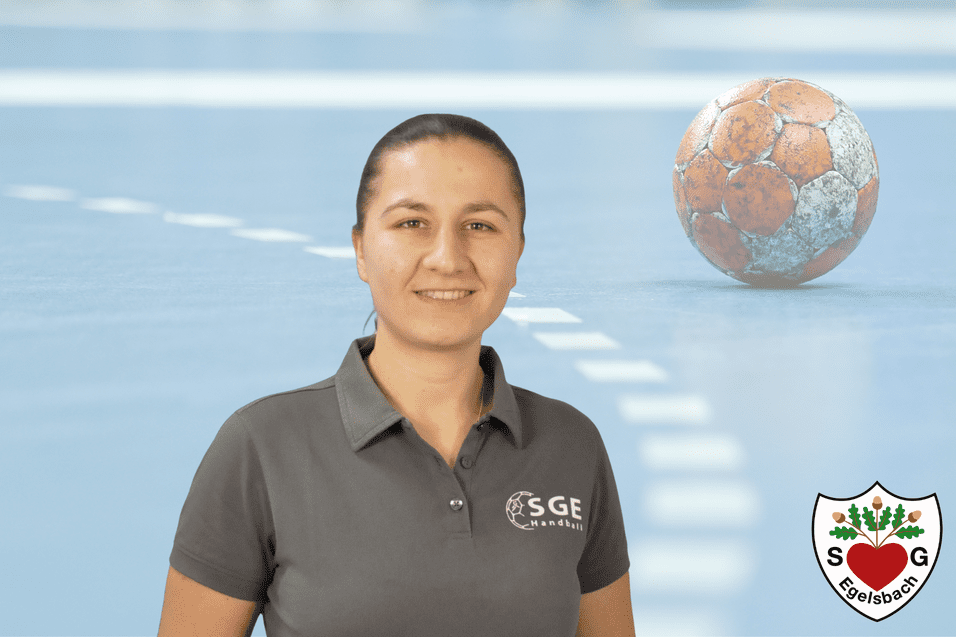Porträt einer lächelnden Frau in grauem Shirt vor einem Handballfeld, ein Handball im Hintergrund.