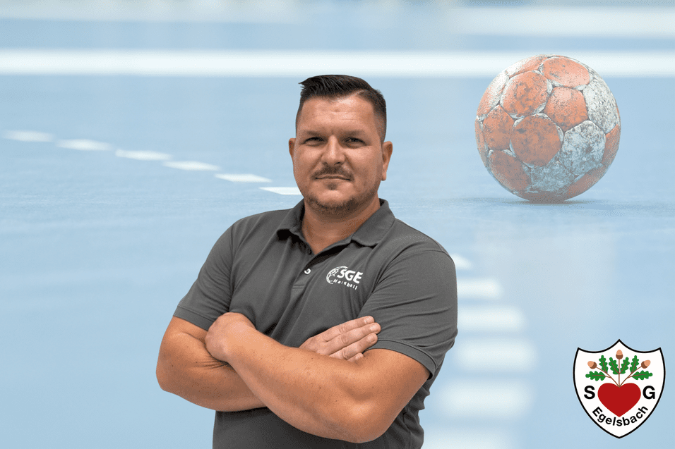 Mann mit verschränkten Armen steht vor einem Handballfeld, im Hintergrund ein Handball und das Logo eines Vereins.