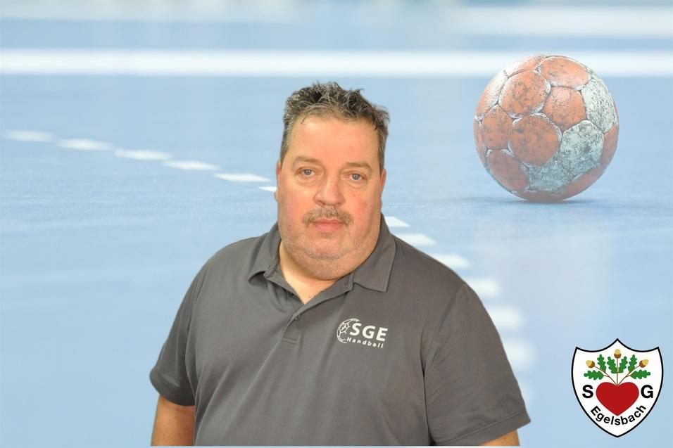 Mann in Sportbekleidung vor einer Handballfläche, im Hintergrund ein Handball auf einem blauen Boden.