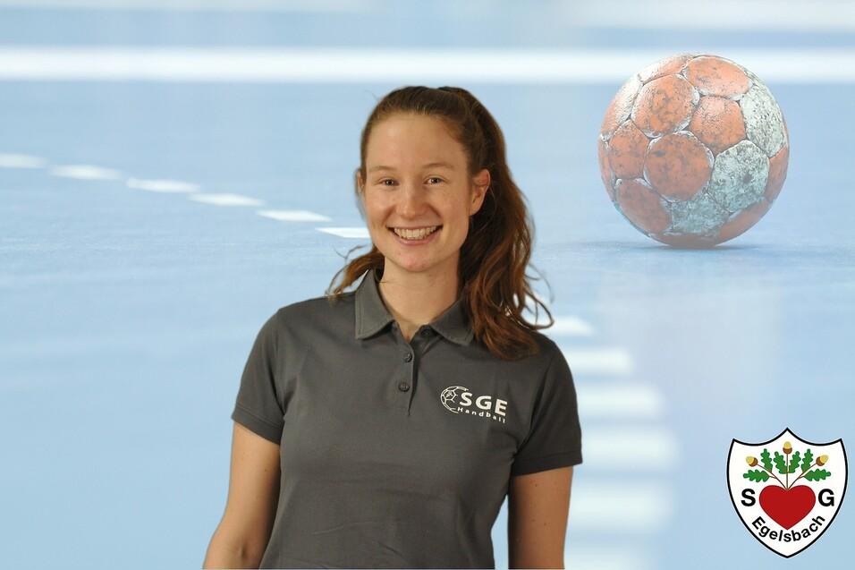 Lächelnde Frau in einem grauen Polo-Shirt vor verschwommenem Handball und Sportfläche. Logo in der Ecke.