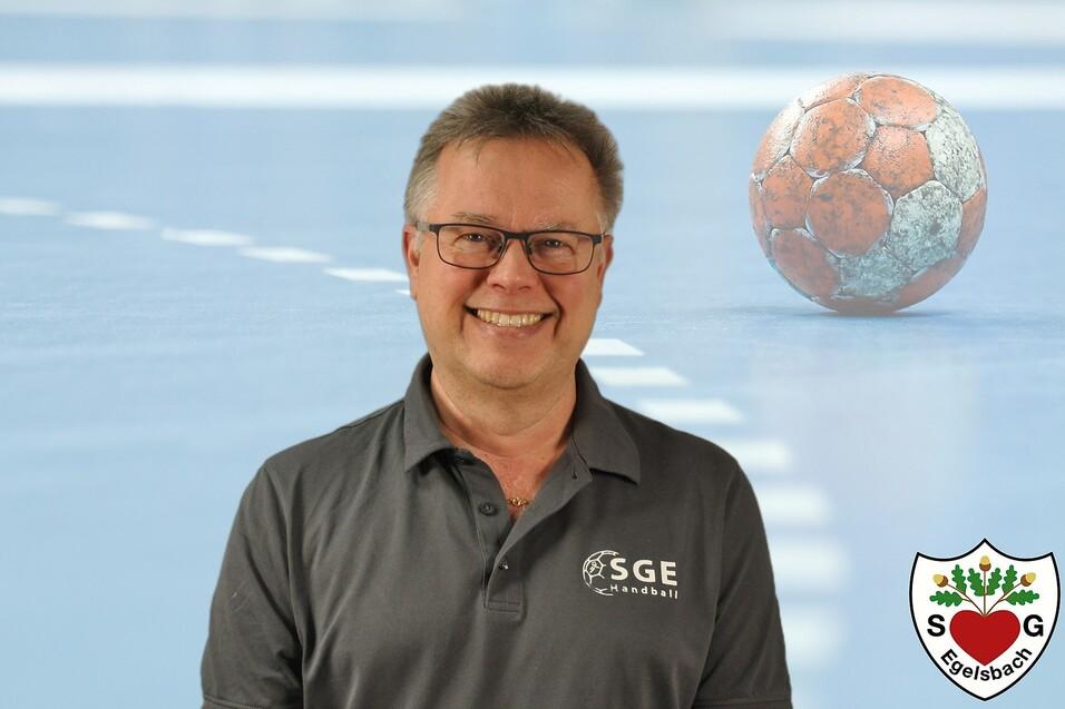 Lächelnder Mann mit Brille trägt ein graues Polo-Shirt vor einer Handballfläche und einem Handball im Hintergrund.