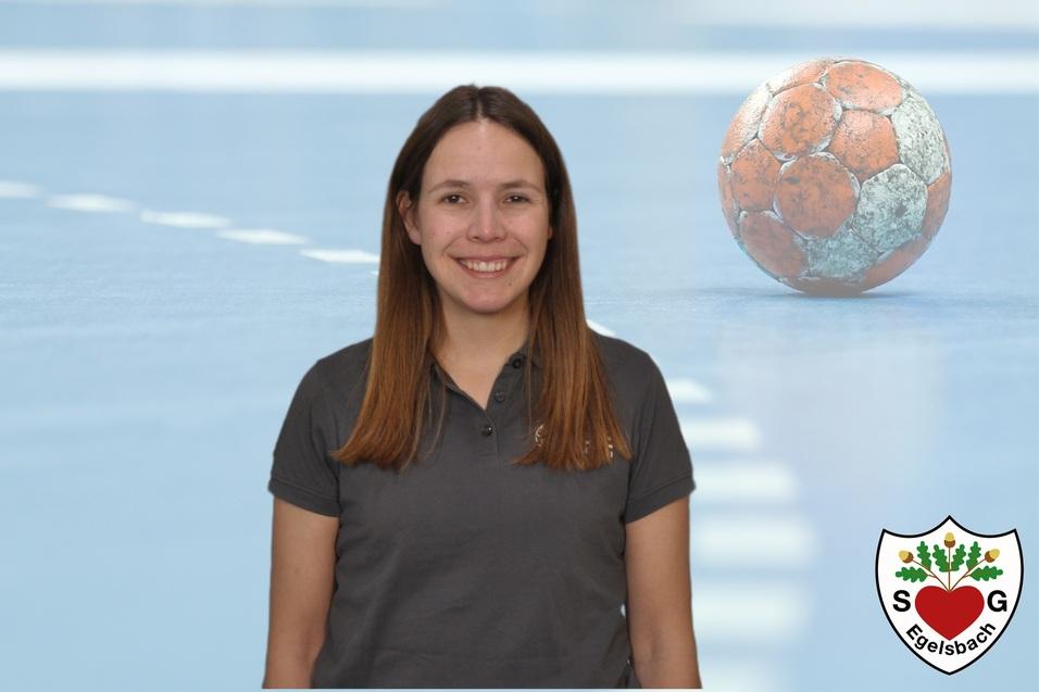 Porträt einer Frau mit braunen Haaren, stehend vor einem Handball und einem blauen Sportboden.