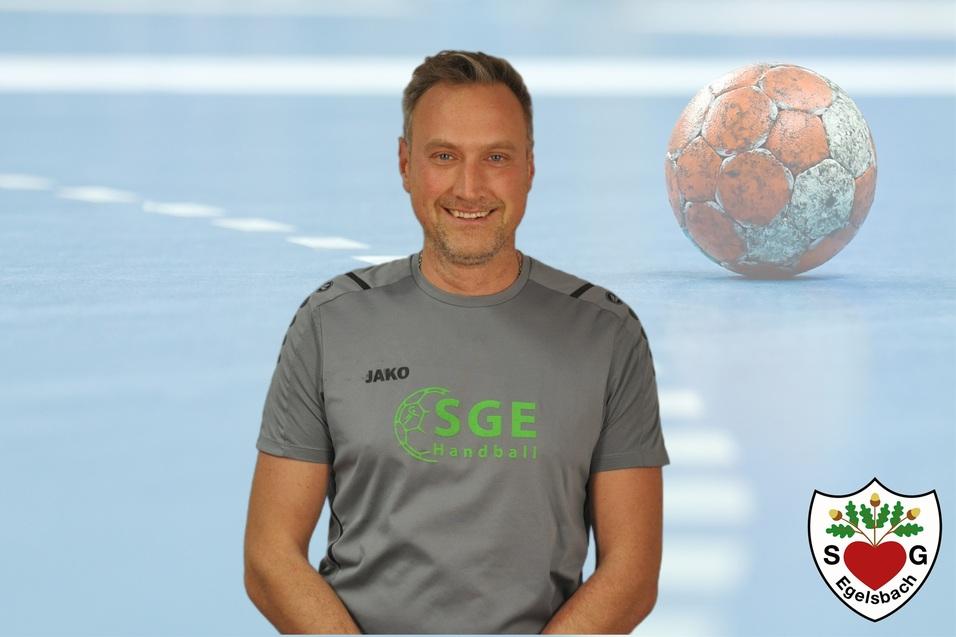 Mann in grauem Sportshirt mit grüner Schrift steht lächelnd vor einer Handball-Spielfeld-Hintergrund mit Ball.