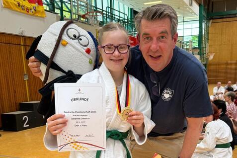 M&auml;dchen in Judogi h&auml;lt Urkunde und Medaille, posiert mit Trainer in Sporthalle vor versammelter Jury.