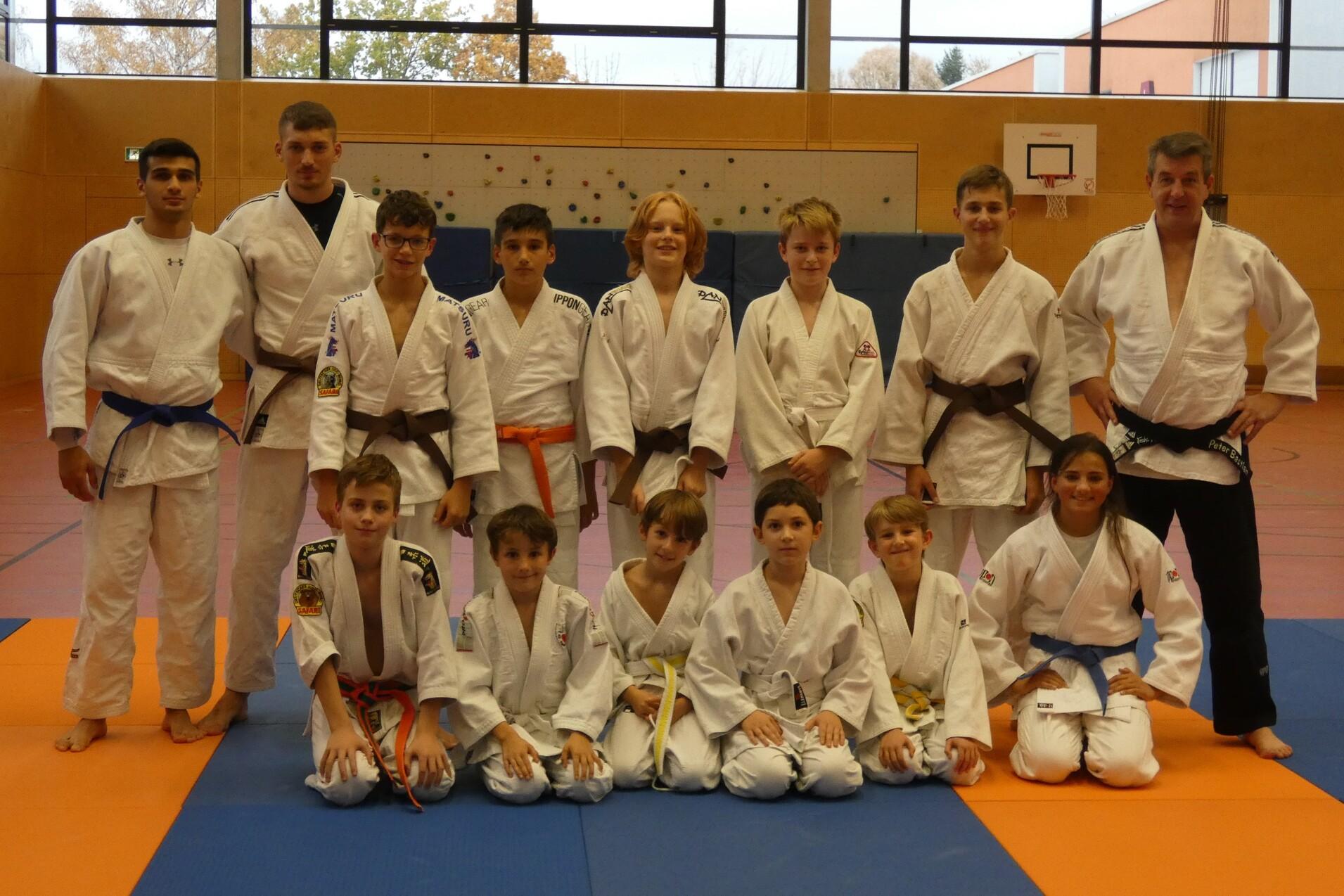 Gruppenshot von 14 Judoka in verschiedenen G&uuml;rtelfarben auf einer Matte in einem Trainingsraum.