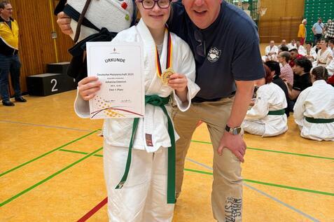 M&auml;dchen in Judogi h&auml;lt Urkunde und Medaille, posiert mit Trainer in Sporthalle vor versammelter Jury.