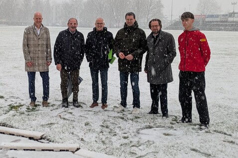 Sechsergruppe von M&auml;nnern in Winterjacken steht im Schneefall auf einem Sportplatz mit gr&uuml;nem Rasen.