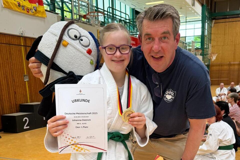 M&auml;dchen in Judogi h&auml;lt Urkunde und Medaille, posiert mit Trainer in Sporthalle vor versammelter Jury.