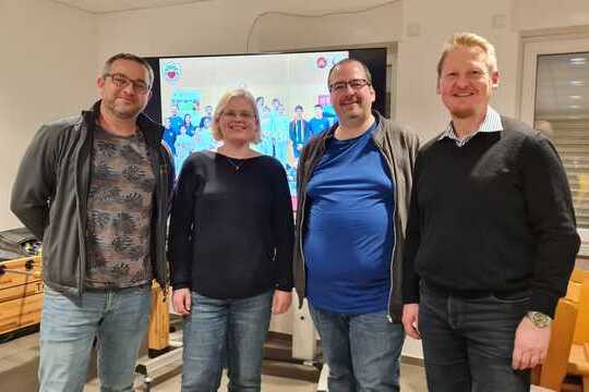 Vier Personen stehen smiling nebeneinander vor einem Bildschirm, der eine Gruppenaufnahme im Hintergrund zeigt.