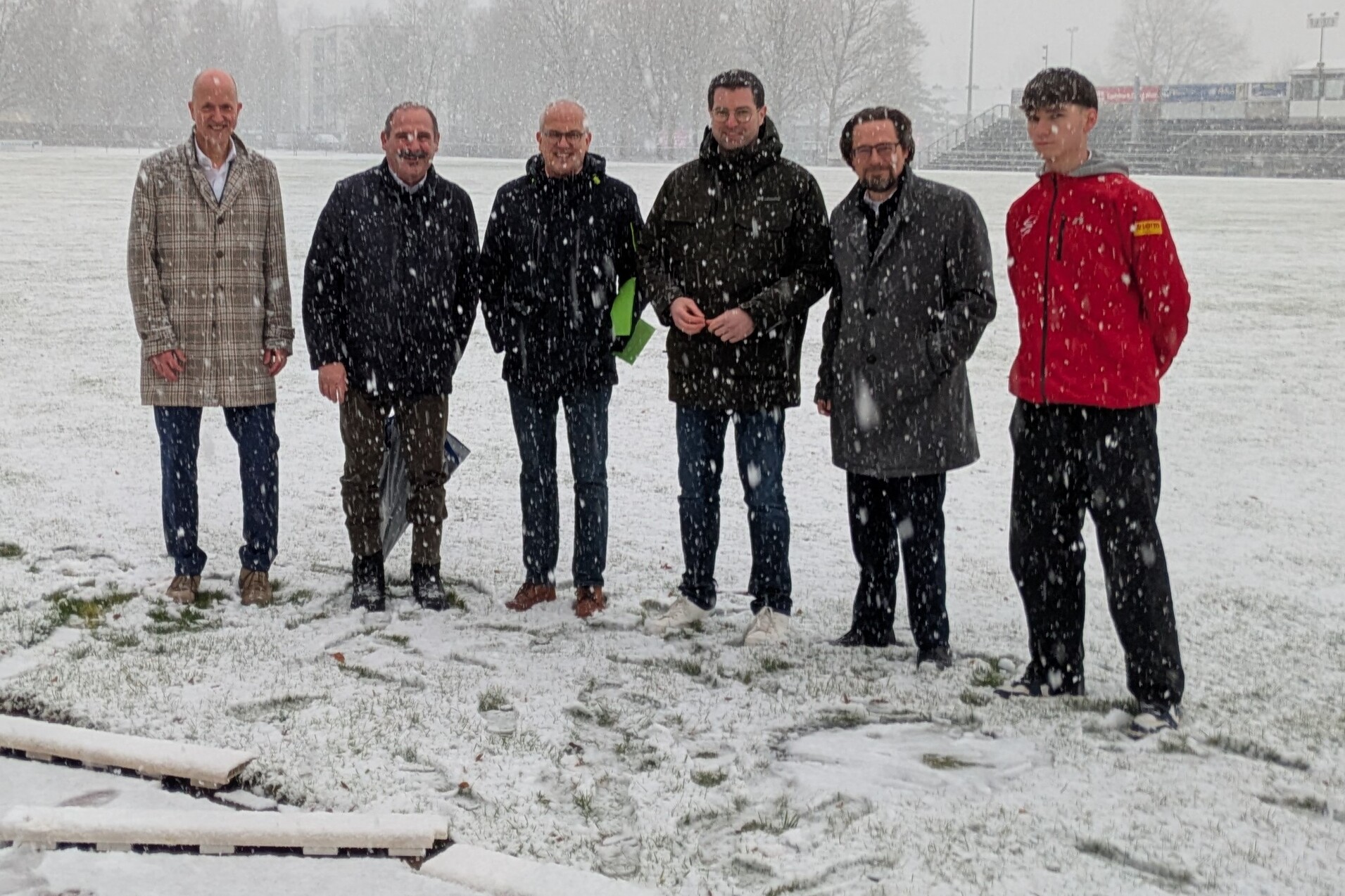 Sechsergruppe von M&auml;nnern in Winterjacken steht im Schneefall auf einem Sportplatz mit gr&uuml;nem Rasen.
