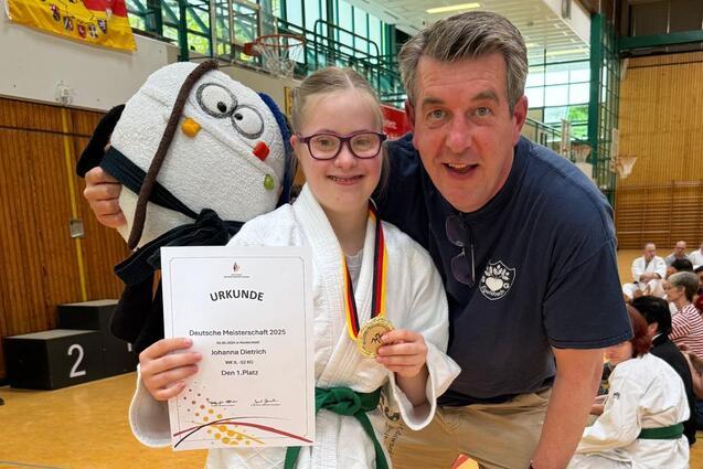 M&auml;dchen in Judogi h&auml;lt Urkunde und Medaille, posiert mit Trainer in Sporthalle vor versammelter Jury.
