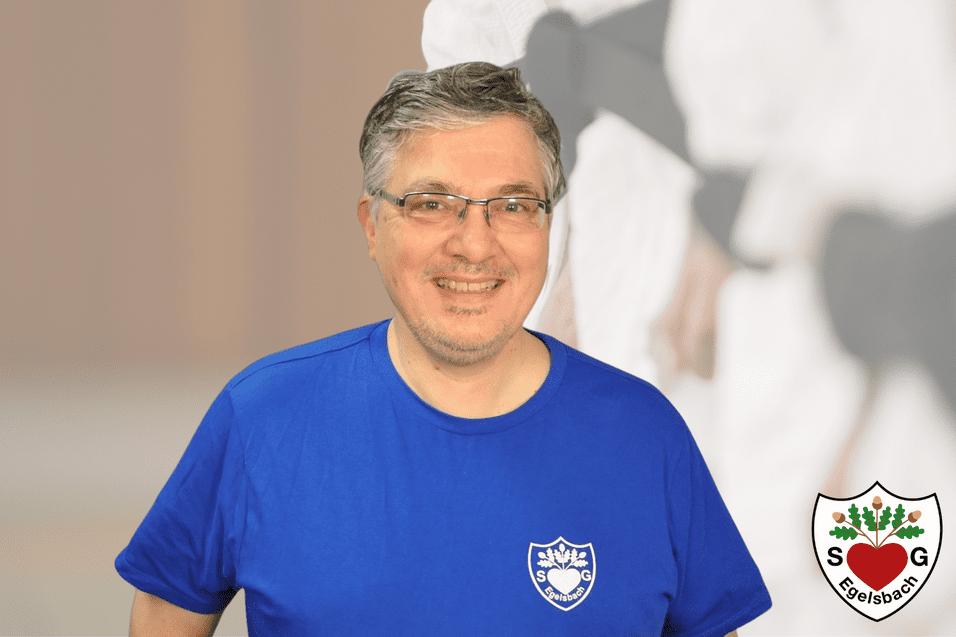 Mann in blauer Sportbekleidung lächelt vor verschwommenem Hintergrund mit einem Sportlogo in der Ecke.