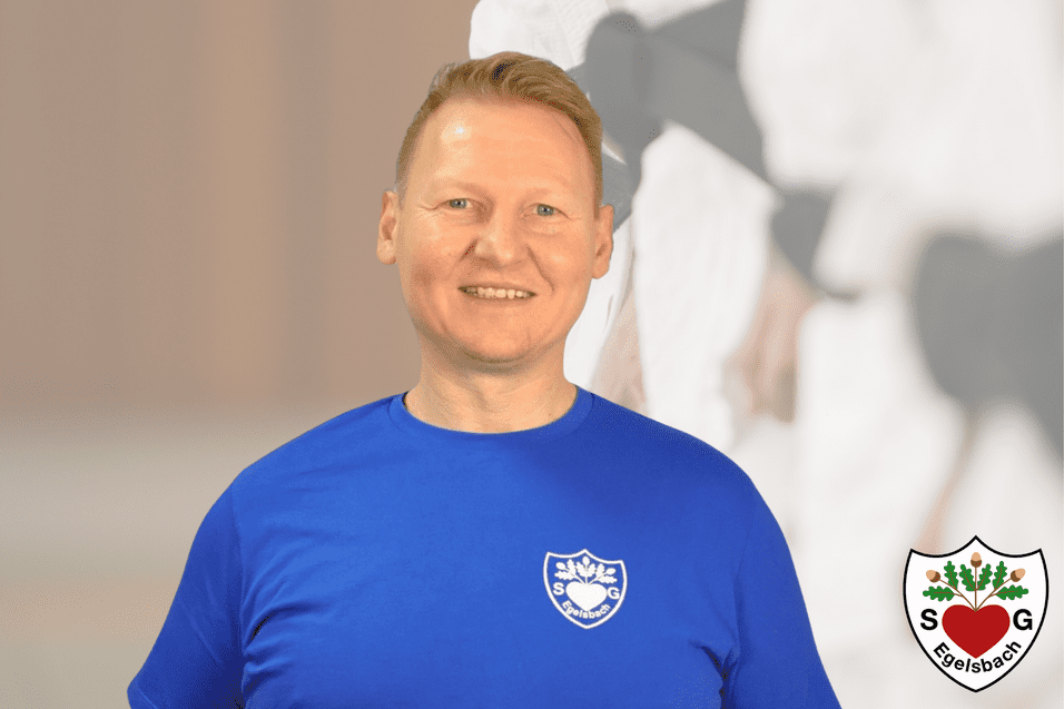 Lächelnder Mann in blauem T-Shirt steht vor unscharfem Hintergrund mit sportlicher Darstellung.