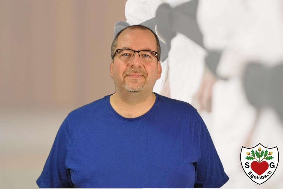 Mann mit Brille und blauer T-Shirt vor unscharfem Hintergrund, auf dem Elemente des Kampfsports zu sehen sind.