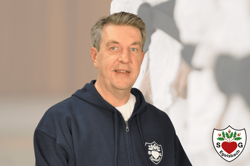 Porträt eines Mannes in einem blauen Sweatshirt mit festem Blick, vor unscharfem Hintergrund einer Sportanlage.