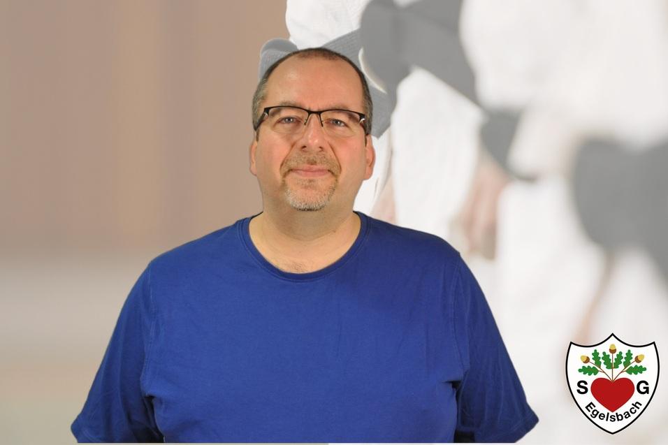 Mann mit Brille und blauer T-Shirt vor unscharfem Hintergrund, auf dem Elemente des Kampfsports zu sehen sind.