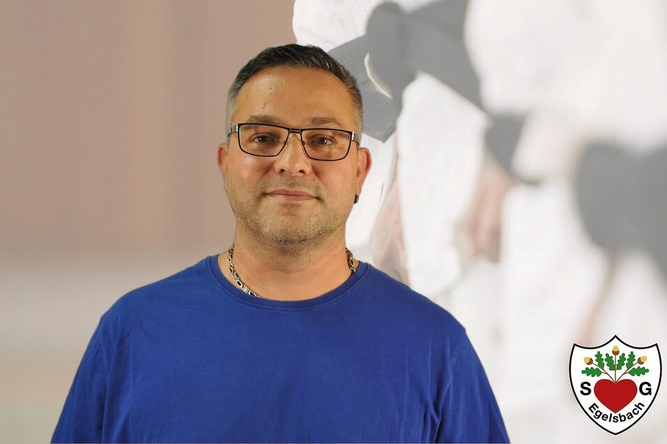 Portrait eines Mannes mit Brille in blauem T-Shirt vor einem verschwommenen Hintergrund mit Kampfsportern.