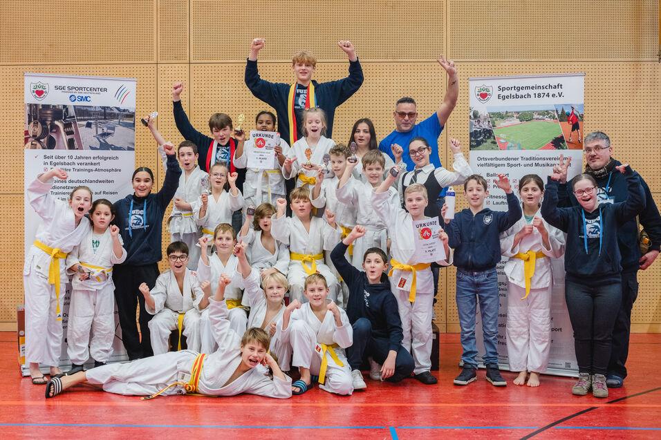 Gruppe von Kindern in Judoanz&uuml;gen mit Medaillen, die vor zwei Bannern in einer Sporthalle feiern.