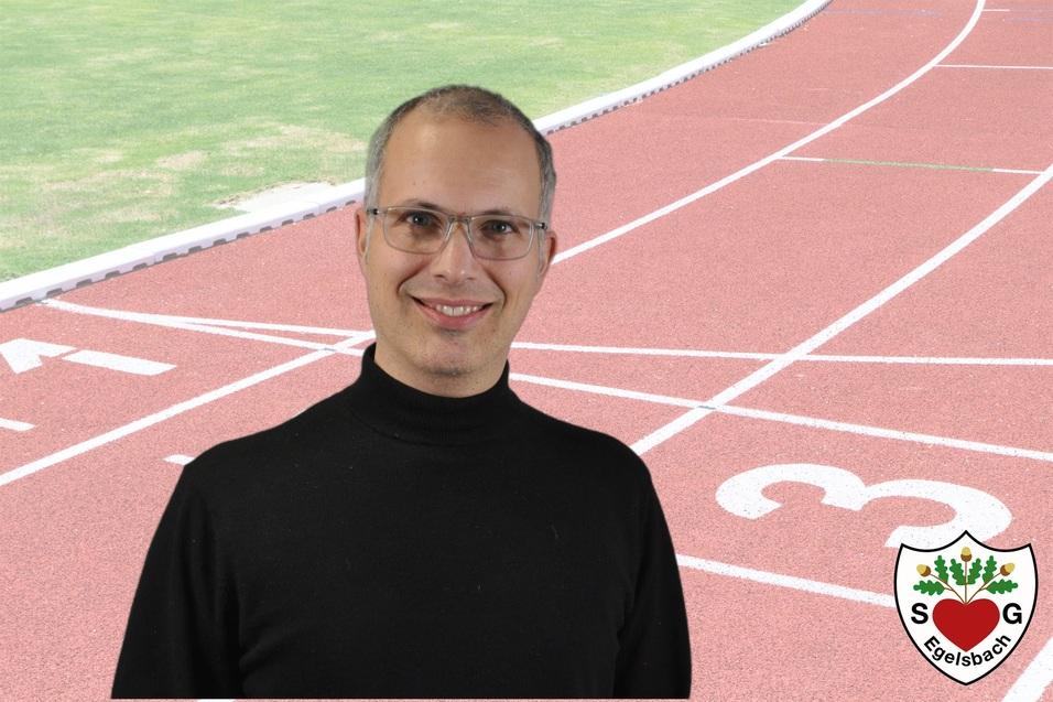Portr&auml;t eines Mannes mit Brille vor einer Laufbahn, die Startlinien und das Vereinslogo zeigt.