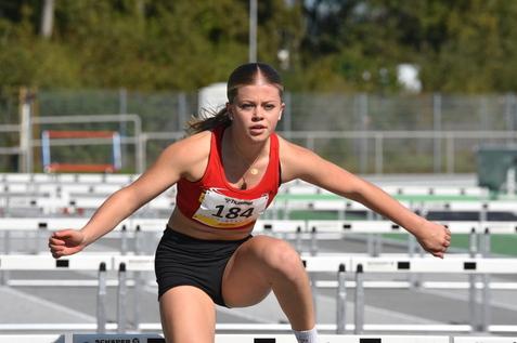 Junge Sportlerin in rot-schwarzer Wettkampfkleidung springt &uuml;ber H&uuml;rden auf einer Laufbahn im Freien.