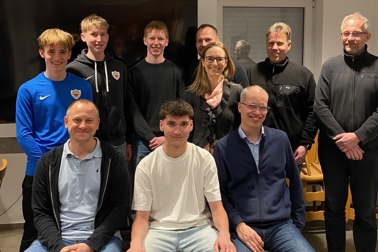 Gruppenfoto mit acht Personen, die verteilt auf St&uuml;hlen stehen und sitzen, vor einem gro&szlig;en Bildschirm.
