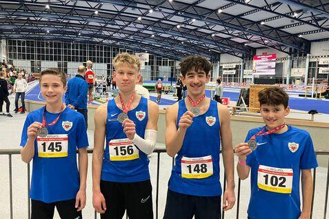 Vier junge Athleten in blauen Trikots mit Medaillen stehen l&auml;chelnd in einer Sporthalle w&auml;hrend eines Wettkampfs.