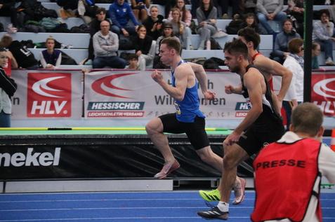 Sprintende Athleten auf einer blauen Laufbahn, Zuschauer im Hintergrund, w&auml;hrend eines Wettkampfs in einer Halle.
