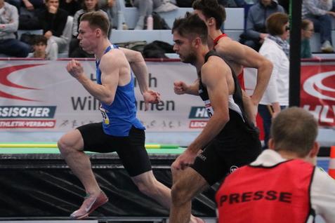Sprintende Athleten auf einer blauen Laufbahn, Zuschauer im Hintergrund, w&auml;hrend eines Wettkampfs in einer Halle.
