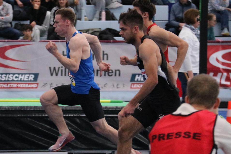 Sprintende Athleten auf einer blauen Laufbahn, Zuschauer im Hintergrund, w&auml;hrend eines Wettkampfs in einer Halle.