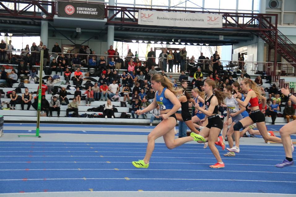 M&auml;dchen sprinten auf einer blauen Leichtathletikbahn w&auml;hrend eines Wettkampfs, Zuschauer sitzen im Hintergrund.