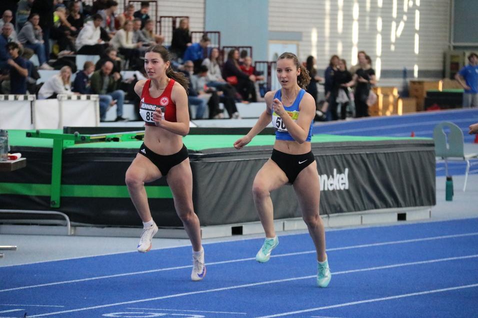 Zwei Athletinnen sprinten auf einer blauen Laufbahn w&auml;hrend eines Wettkampfs in einer Sporthalle.
