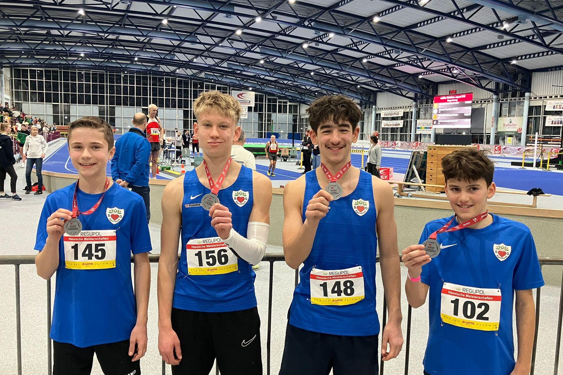 Vier junge Athleten in blauen Trikots mit Medaillen stehen l&auml;chelnd in einer Sporthalle w&auml;hrend eines Wettkampfs.