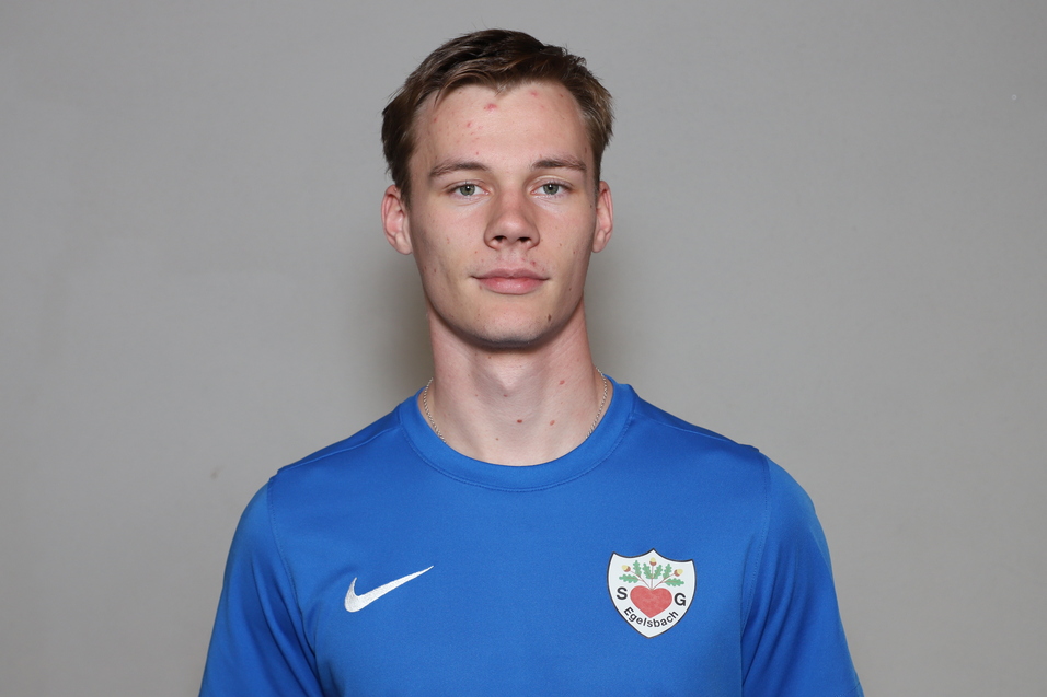 Junger Mann mit kurzem Haar, tr&auml;gt ein blaues Sportshirt und steht vor einem neutralen grauen Hintergrund.