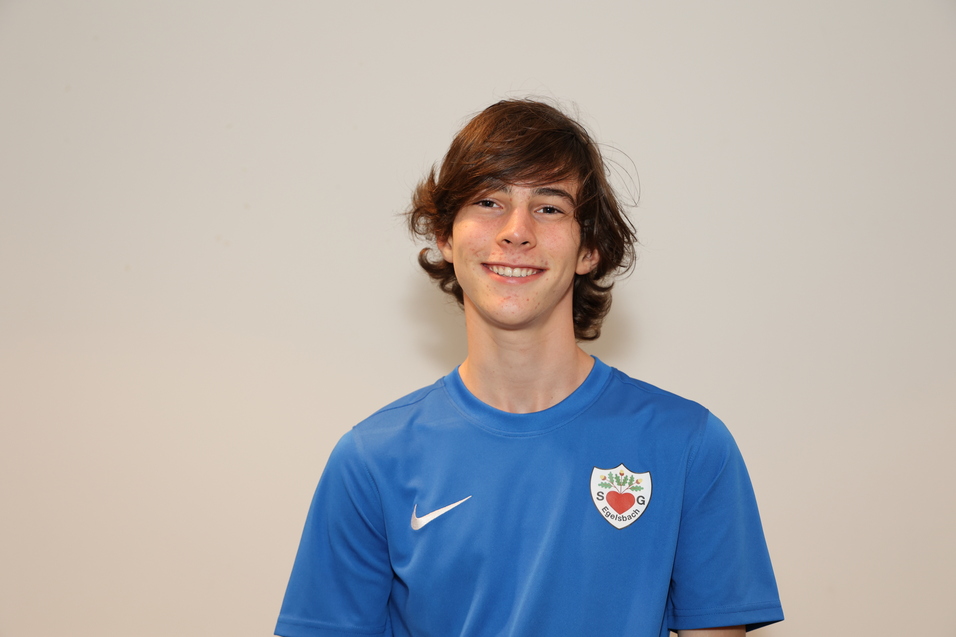 Junger Mann mit lockigem Haar tr&auml;gt ein blaues Fu&szlig;balltrikot und l&auml;chelt vor einem neutralen Hintergrund.